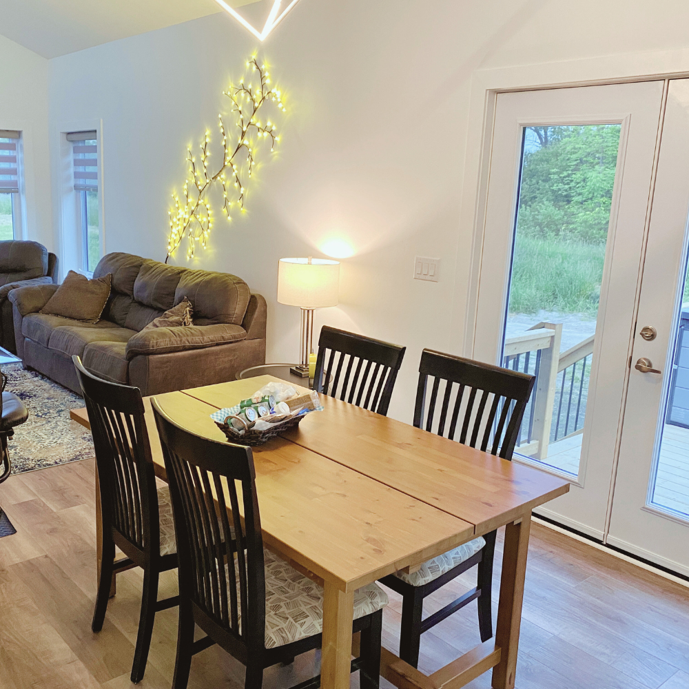 Bright and cozy dining and living area at Port Albert Paradise featuring a wooden dining table with six cushioned chairs, a comfy brown sectional sofa, and a unique wall decoration made of glowing branch-style lights.